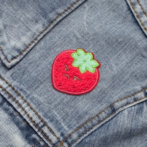 Può includere: Una toppa ricamata a forma di fragola rossa con gambo e foglie verdi. La fragola ha un'espressione arrabbiata da cartone animato.