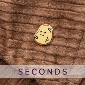 Op de afbeelding: Een goudomrande pin met een lichtgeel, aardappelvormig ontwerp en zwarte gezichtskenmerken. De pin is geplaatst tegen een bruine, corduroy-achtige achtergrond. Het woord "SECONDS" wordt in een paarse banner onderaan weergegeven.