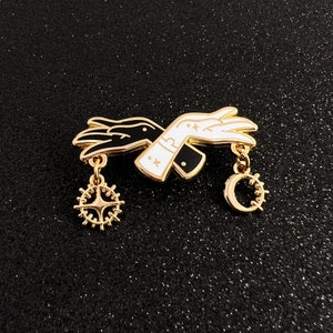 Op de afbeelding: Gouden emaille pin met twee handen, één zwart en één wit, die elkaar vasthouden. De pin heeft twee hangende bedels, een halve maan en een ster.