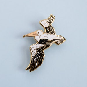 May include: A gold-coloured enamel pin of a white pelican with black wings in flight. The pelican has a long, thin beak and a small, round head.