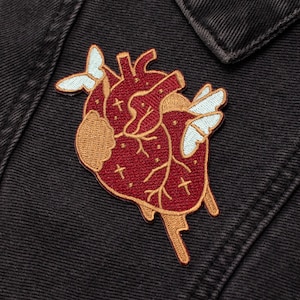 Könnte beinhalten: Ein rotes anatomisches Herz mit goldenen Nähten und zwei weißen Schmetterlingen auf einer schwarzen Jeansjacke. Das Herz hat einen tropfenden Effekt und kleine Kreuze.
