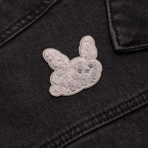 May include: A grey, embroidered bunny patch with a fluffy texture and a silver outline. The patch is attached to a dark denim jacket, showing the details of the bunny's face and ears. The patch is a cute accessory.
