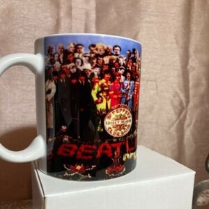 May include: White ceramic mug featuring a colourful image of the Beatles band members on the front. The image includes the band's name and the album title "Sgt. Pepper's Lonely Hearts Club Band".