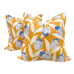 May include: Two square throw pillows with a vibrant yellow background and a repeating floral pattern. The pattern features white leaves and blue and white tulip-like flowers. The pillows are a decorative home accent.