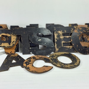 May include: A set of vintage wooden letters, painted black with gold accents, spelling out the word "ARTISTIC". The letters are worn and have a distressed look, giving them a unique and rustic charm.