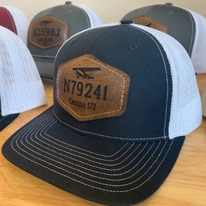 May include: A collection of five trucker hats with a white mesh back and a brown leather patch on the front. The patches feature a small airplane graphic and the text "N3598J Cessna 150" and "N79241 Cessna 172".