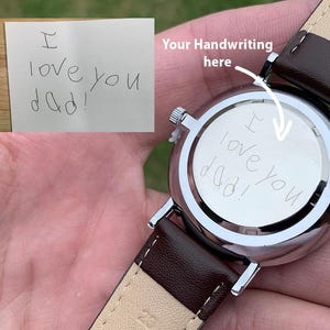 May include: A silver wristwatch with a brown leather strap. The watch face has the handwritten message "I love you dad!" engraved on the back. The watch is held in a person's hand.