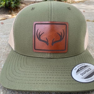 Pu&ograve; includere: Un cappello da camionista verde e beige con una toppa in pelle marrone con un design a corna di cervo. Il cappello ha una parte posteriore in rete e un'etichetta nera "The Authentic Yupoong Snapback".