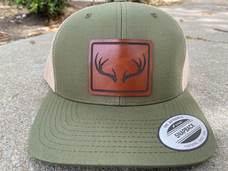 Pu&ograve; includere: Un cappello da camionista verde e beige con una toppa in pelle marrone con un design a corna di cervo. Il cappello ha una parte posteriore in rete e un'etichetta nera "The Authentic Yupoong Snapback".