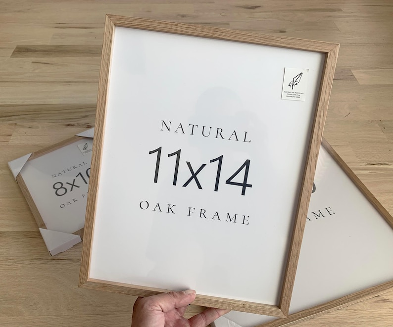 A person is holding a white picture frame with the text "NATURAL 11x14 OAK FRAME" on it.