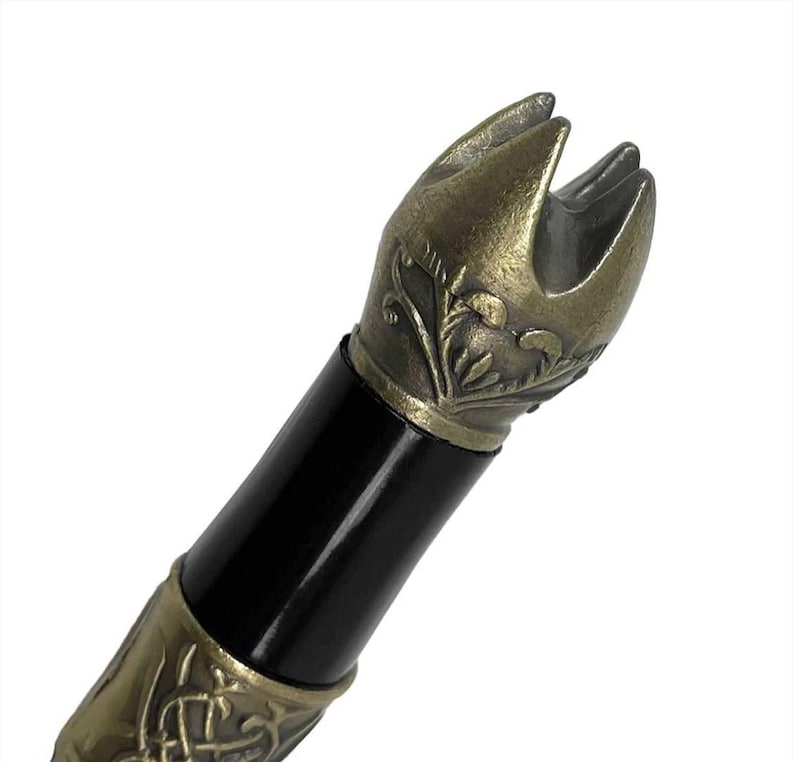 May include: Close-up of the ornate, gold-toned pommel of a sword. The pommel is detailed with intricate floral designs and has a pointed, crown-like top.