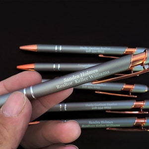 May include: A close-up of a gray metal pen with a rose gold accent. The pen is engraved with the text "Sandra Holmes Realtor Keller Williams" and a phone number.