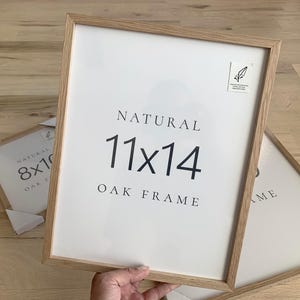 A person is holding a white picture frame with the text "NATURAL 11x14 OAK FRAME" on it.
