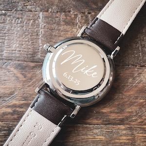 May include: A silver wristwatch with a brown and cream leather band. The watch face is engraved with the name "Mike" and the date "6.13.25". The watch is set against a wooden background.