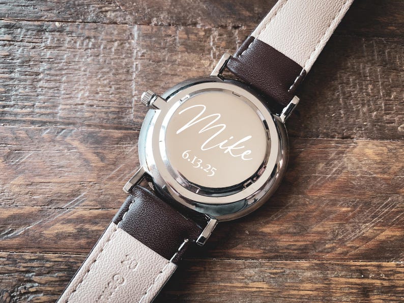 May include: A silver wristwatch with a brown and cream leather band. The watch face is engraved with the name "Mike" and the date "6.13.25". The watch is set against a wooden background.
