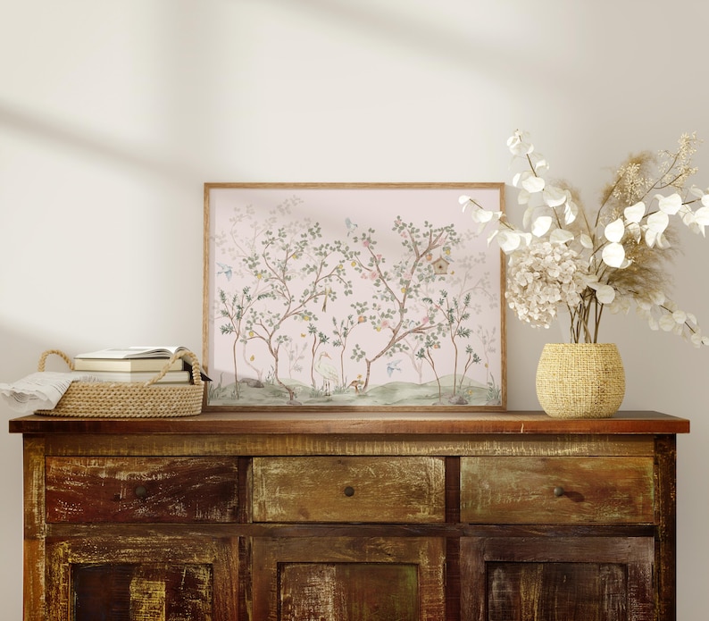a wooden cabinet with a framed picture of trees and flowers on top of it, along with a woven basket and a vase of white flowers.
