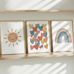 May include: Three framed prints with a minimalist design. The first print features a smiling sun with yellow rays. The second print features a cluster of colorful hearts. The third print features a rainbow with a pink, orange, and blue color scheme.