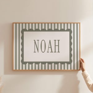 A person is holding a framed picture with the name "Noah" on it, which is hanging on a wall.