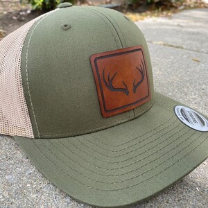 Pu&ograve; includere: Un cappello da camionista verde e beige con una toppa in pelle marrone che presenta un design a corna di cervo. Il cappello &egrave; realizzato dal marchio "Swagman".