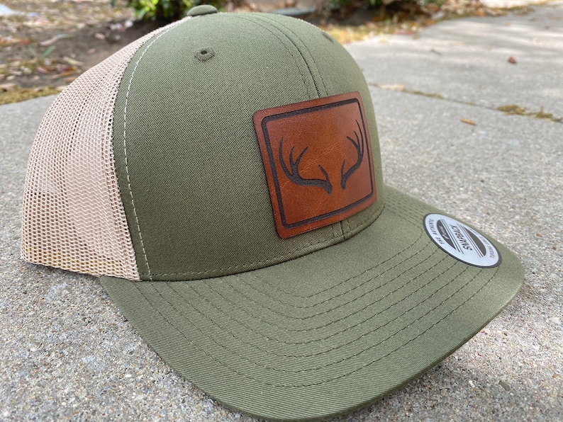 Pu&ograve; includere: Un cappello da camionista verde e beige con una toppa in pelle marrone che presenta un design a corna di cervo. Il cappello &egrave; realizzato dal marchio "Swagman".
