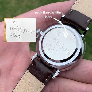May include: A silver wristwatch with a brown leather strap. The watch face has the handwritten message "I love you dad!" engraved on the back. A small note with the same message is also visible.