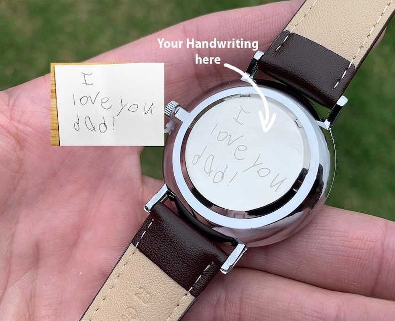 Puede incluir: Un reloj de pulsera plateado con una correa de cuero marr&oacute;n. La parte trasera de la esfera del reloj tiene el mensaje manuscrito "I love you dad!". Tambi&eacute;n es visible una peque&ntilde;a nota con el mismo mensaje.