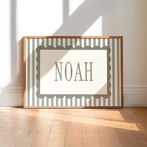 A framed picture with the word "NOAH" displayed on it, placed on a wooden floor against a white wall.