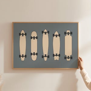 A person is holding up a framed artwork that features five skateboard designs on a blue background.