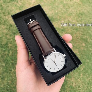 May include: A minimalist wristwatch with a white face and silver-tone case, featuring a brown leather band. The watch is presented in a black gift box. The text "Gift Box Included" is visible.