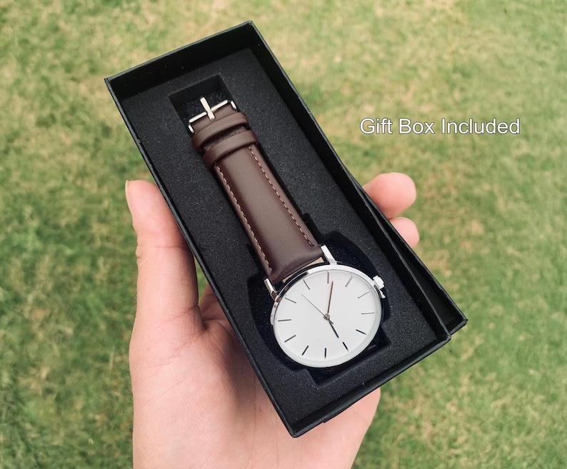 May include: A minimalist wristwatch with a white face and silver-tone case, featuring a brown leather band. The watch is presented in a black gift box. The text "Gift Box Included" is visible.