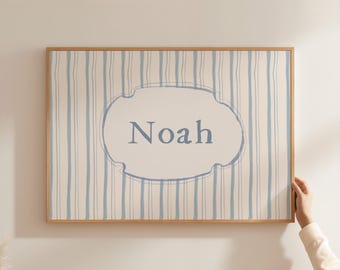 May include: A framed wall art print featuring the name "Noah" in blue script within a light blue and white striped design. The artwork has a light wood frame and is held by a person's hand.