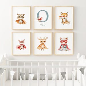 May include: Six framed prints featuring watercolor illustrations of animals wearing superhero capes and masks. The animals include a raccoon, fox, deer, bear, and a bunny. The prints are arranged in a grid pattern on a white wall above a white crib.