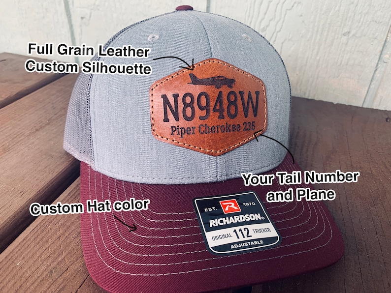 May include: A gray and burgundy trucker hat with a brown leather patch featuring the tail number "N8948W" and the text "Piper Cherokee 235". The hat is adjustable and has the Richardson logo on the back.