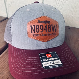 May include: A gray and burgundy trucker hat with a brown leather patch that says "N8948W Piper Cherokee 235". The hat also has a Richardson 112 Trucker label.