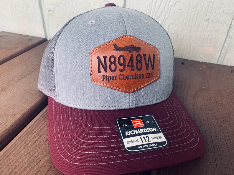 May include: A gray and burgundy trucker hat with a brown leather patch that says "N8948W Piper Cherokee 235". The hat also has a Richardson 112 Trucker label.