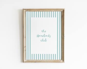 May include: A framed print with a light wood frame. The print has a white background with a light blue striped border. The words "the Homebody club" are written in a light blue script font in the center of the print.