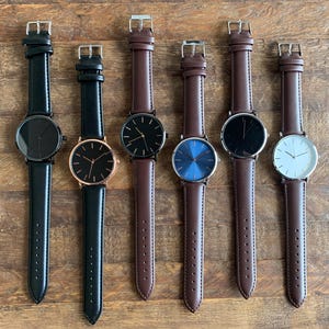 May include: Six wristwatches with leather straps are displayed on a wooden surface. The watches have round faces in black, blue, and white, with gold and silver-toned cases. The watch straps are black and brown.