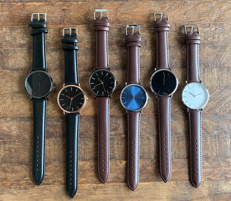 May include: Six wristwatches with leather straps are displayed on a wooden surface. The watches have round faces in black, blue, and white, with gold and silver-toned cases. The watch straps are black and brown.