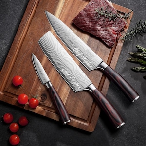 May include: A set of three stainless steel kitchen knives with wooden handles, resting on a wooden cutting board. The blades have a unique pattern and the text "Chef Stephen". Fresh tomatoes and a piece of raw meat are also on the board.