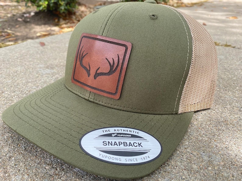 Pu&ograve; includere: Un cappello da camionista verde e beige con una toppa in pelle marrone che presenta un design a corna di cervo. Il cappello ha una parte posteriore in rete e un'etichetta che recita "The Authentic Yupoong Snapback Yupoong Since 1974".