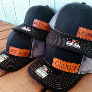Peut inclure: Quatre casquettes de camionneur noires et grises avec des patchs en cuir marron. Les patchs portent les mots "Groom", "Best Man" et "Groomsman".
