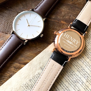 May include: Two wristwatches with brown and black leather bands. One watch has a white face with gold hands and the other has a rose gold face with the inscription "HENRY GROOMSMAN 10.10.23".