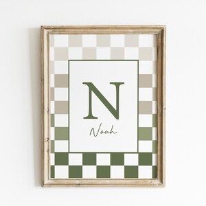 May include: Framed wall art featuring a checkerboard pattern in shades of green and beige. The artwork includes a central white rectangle with the letter "N" and the name "Noah" in a script font. The frame has a light wood finish.