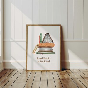 A framed print with a stack of books and the text "Read Books & Be Kind" is displayed on a wooden floor against a white wall.