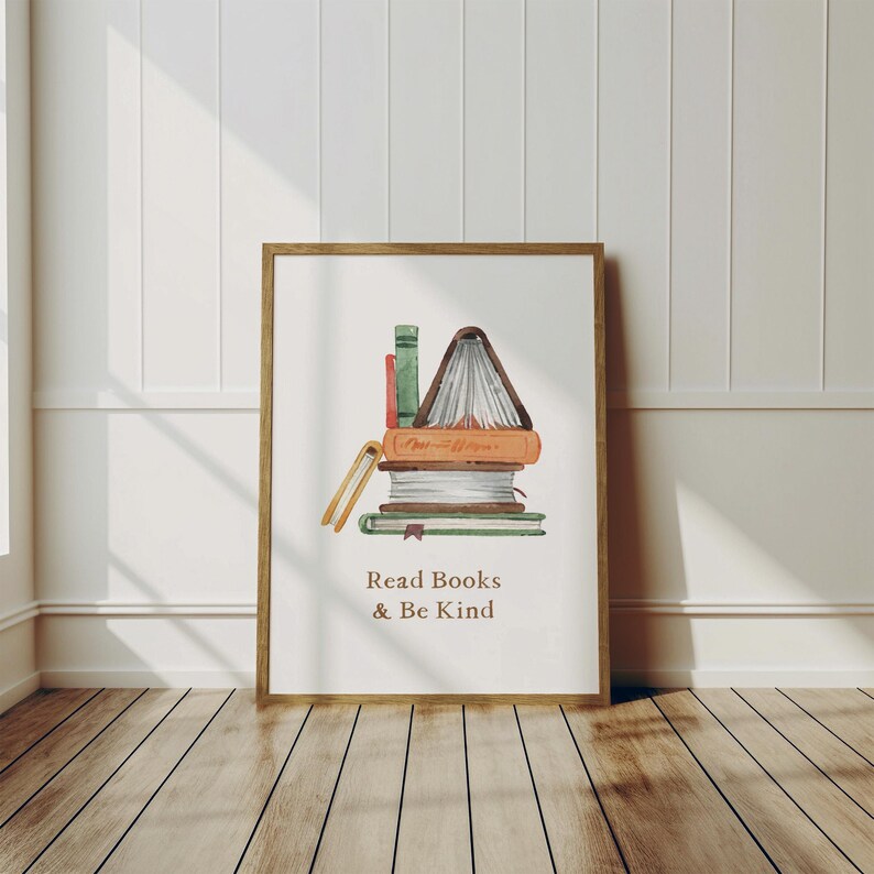 A framed print with a stack of books and the text "Read Books & Be Kind" is displayed on a wooden floor against a white wall.