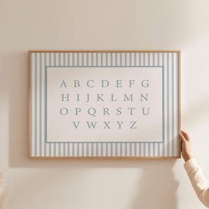 May include: A framed alphabet print with a light blue and white striped border. The center features the letters A through Z in a soft gray font. The frame is a light wood color.