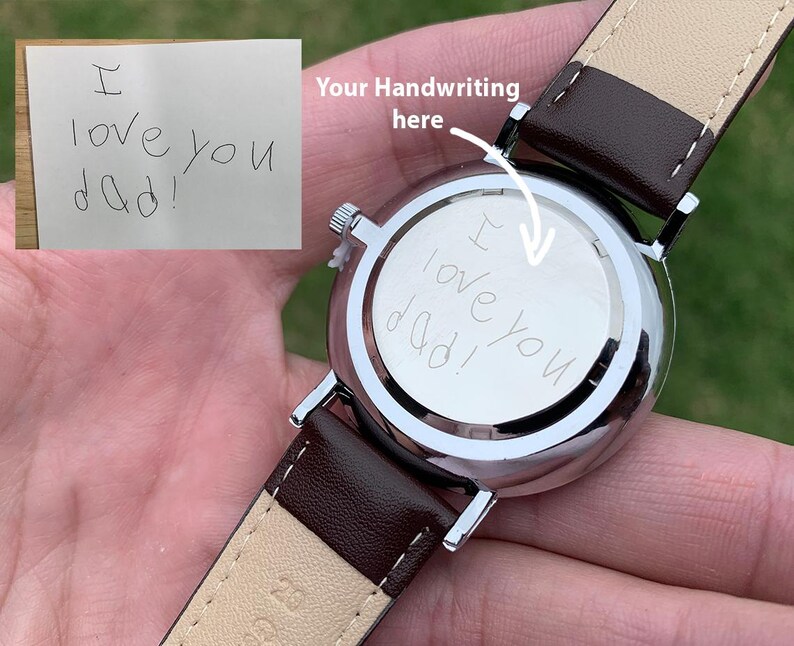 May include: A silver wristwatch with a brown leather strap. The watch face has the handwritten message "I love you dad!" engraved on the back. The watch is held in a person's hand.
