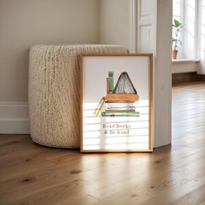 A framed print with an illustration of stacked books and the text "Read Books & Be Kind" is displayed on a wooden floor next to a round white ottoman.