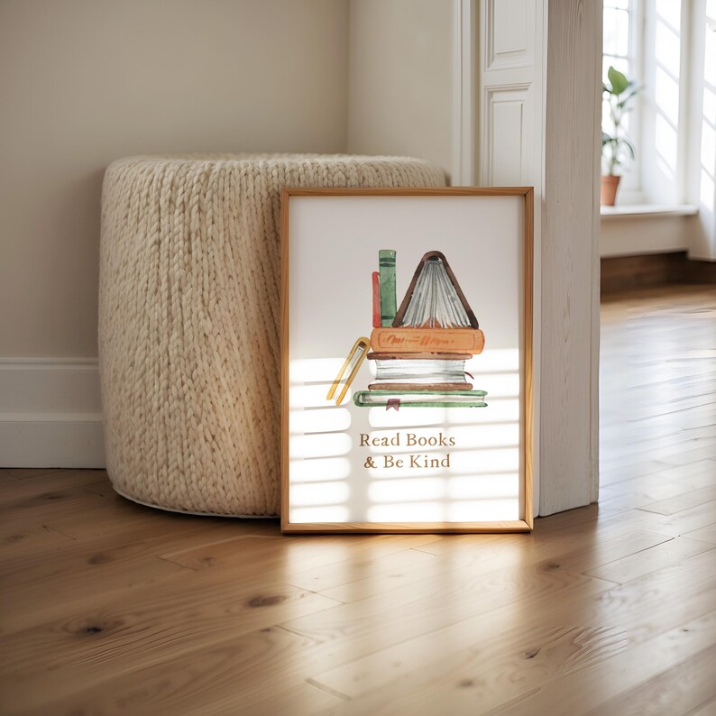 A framed print with an illustration of stacked books and the text "Read Books & Be Kind" is displayed on a wooden floor next to a round white ottoman.