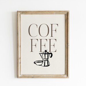 May include: Framed print with the word "COFFEE" in brown capital letters. Below the word is a black and white illustration of a coffee maker and a cup. The frame is a light brown color.